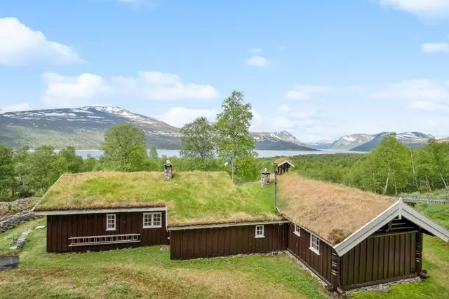 Hytta er perfekt tilpasset tomten og landskapet, med tradisjonelt torvtak, mørk kledning og hvit staffasje.