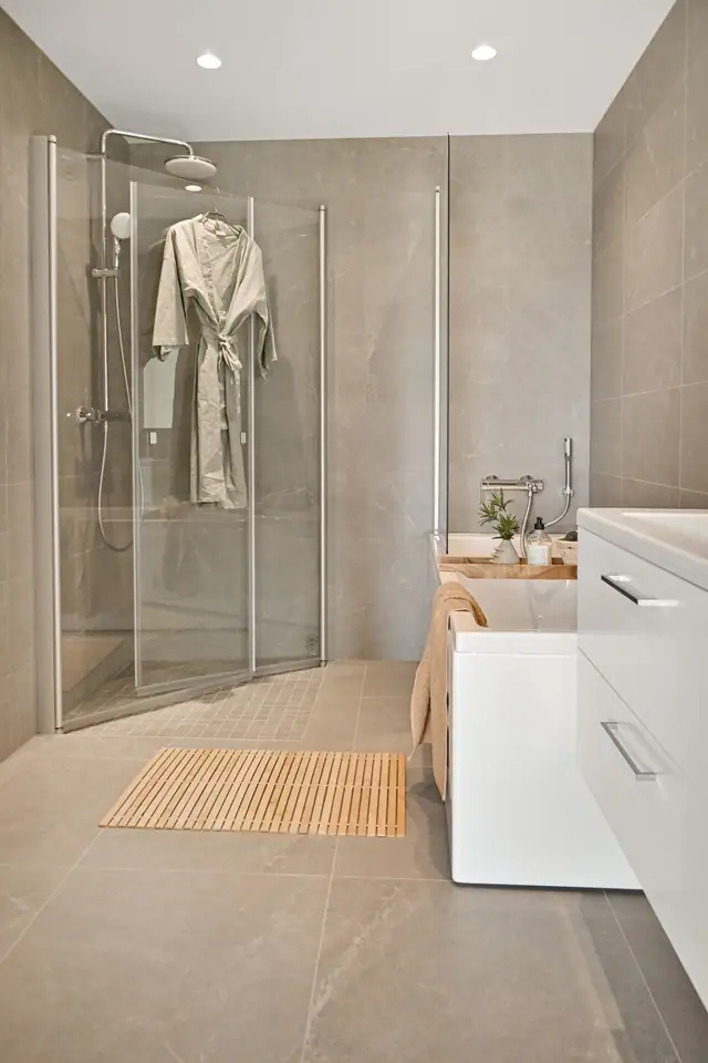 Badet leveres med lysegrå veggfliser, hvit baderomsinnredning, speil med belysning og dusjhjørne.